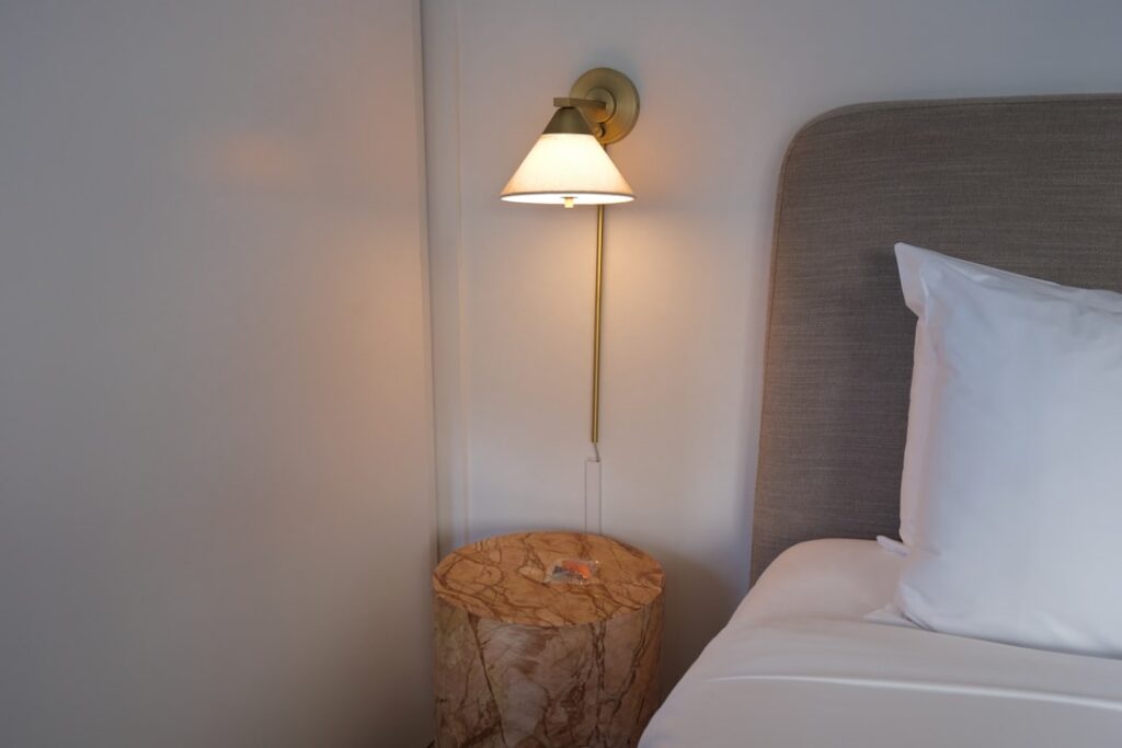 Bedside nook with wall lamp, rustic stump table and crisp white pillows in a small guest space